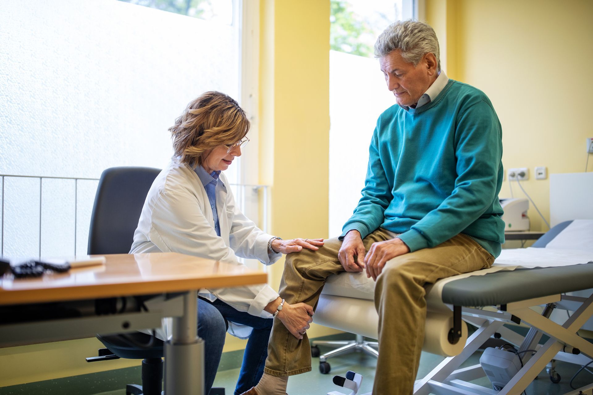 Doctor in white coat examines knee of seated older man wearing a blue sweater in an exam room.