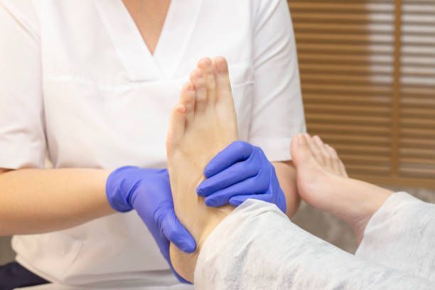 A person in a white uniform examines a patient's foot, wearing blue gloves. Indoor setting.
