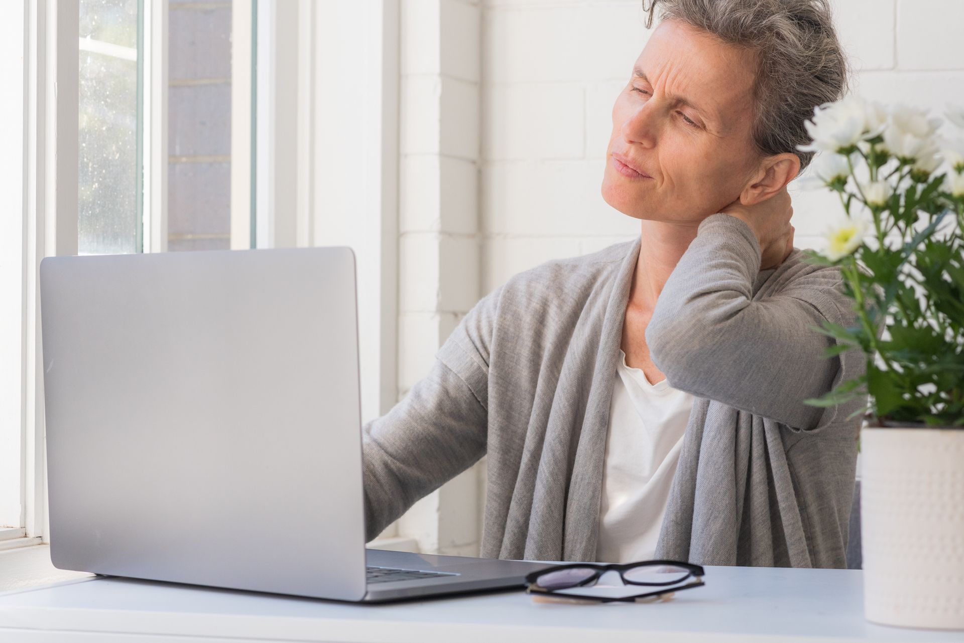 Woman with gray hair, seated at a laptop, touching her neck in pain, with glasses on the table.