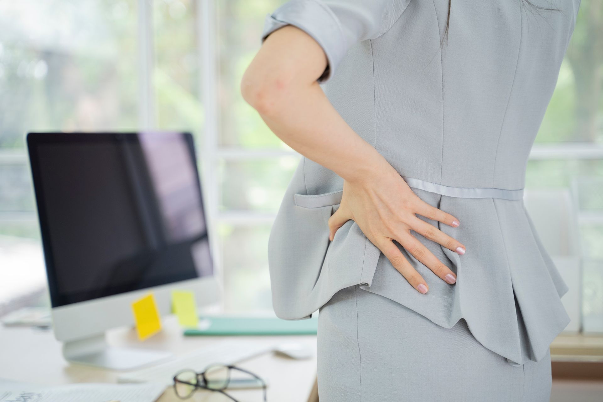 Woman in gray suit holding her lower back, likely in pain, at an office desk with a computer.