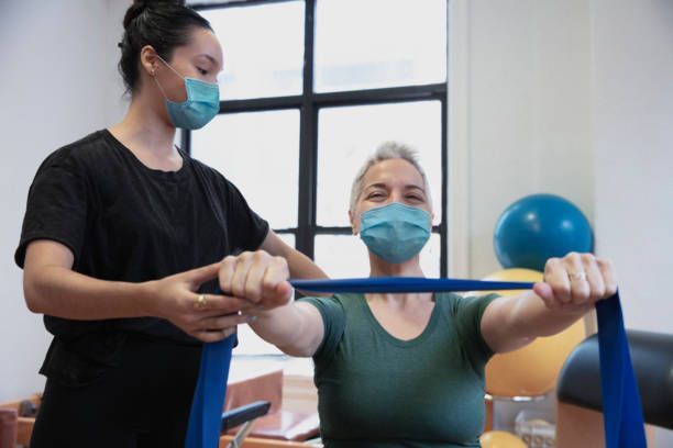A masked instructor assists a woman with a blue resistance band. They are indoors, possibly a studio.