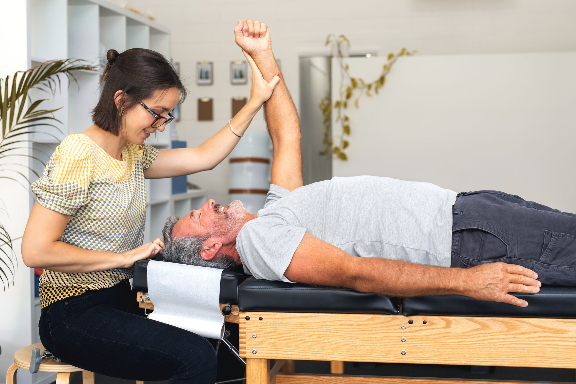 Woman assisting a man on a medical table. She raises his arm as he looks upward, smiling.
