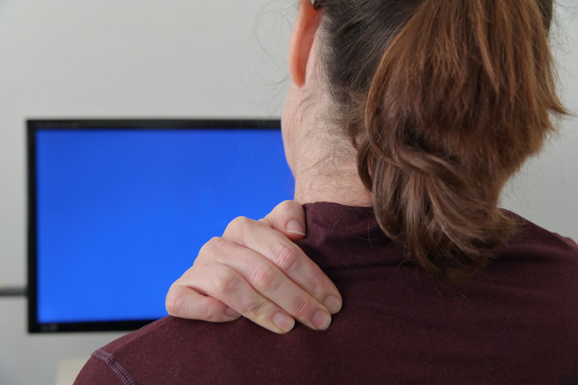 Woman with reddish-brown hair holds her right shoulder in pain while sitting at a computer with a blue screen.