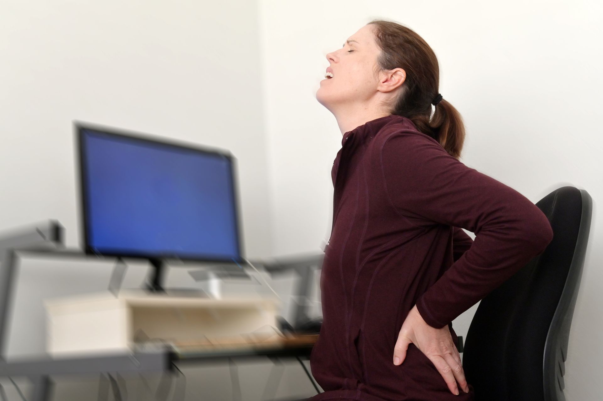 Woman at a desk, clutching her lower back in pain while sitting in an office chair, face scrunched up.