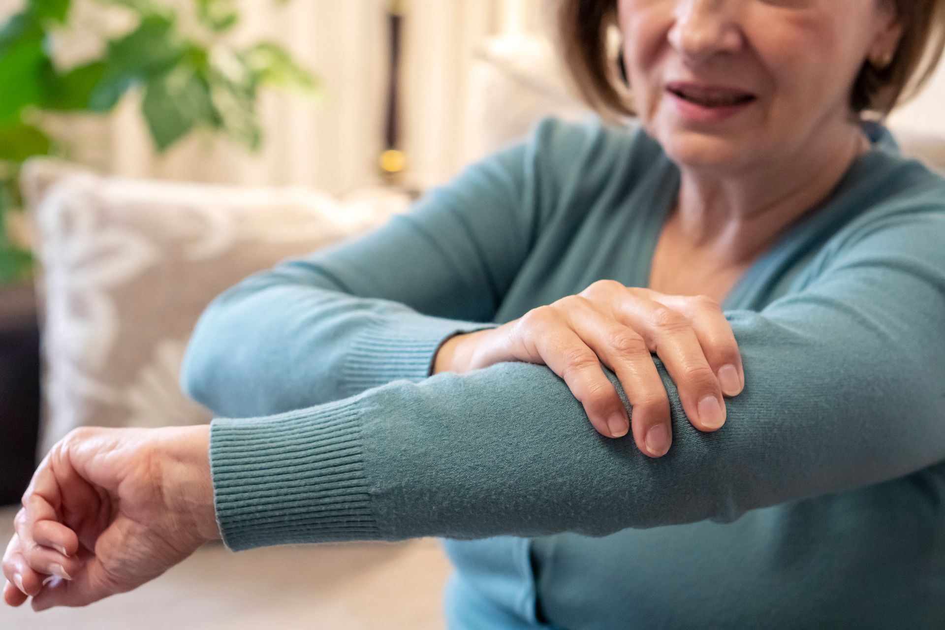 An older woman with fair skin holds her left arm, likely in pain. She wears a blue sweater in a brightly lit room.