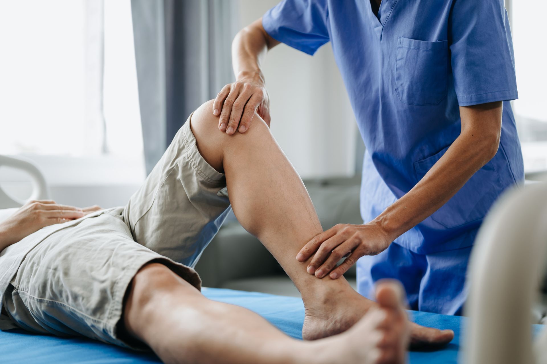 A medical professional in blue scrubs examines a patient's leg, likely checking the knee and ankle on a hospital bed.