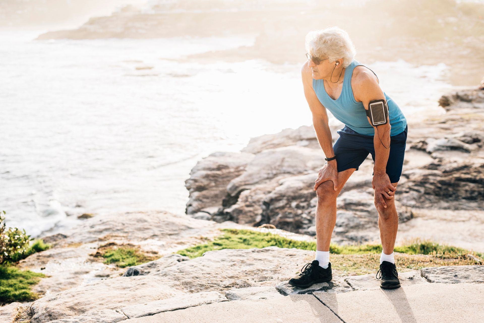Older person in blue tank top and shorts, hands on knees, catching breath after a run near the ocean.