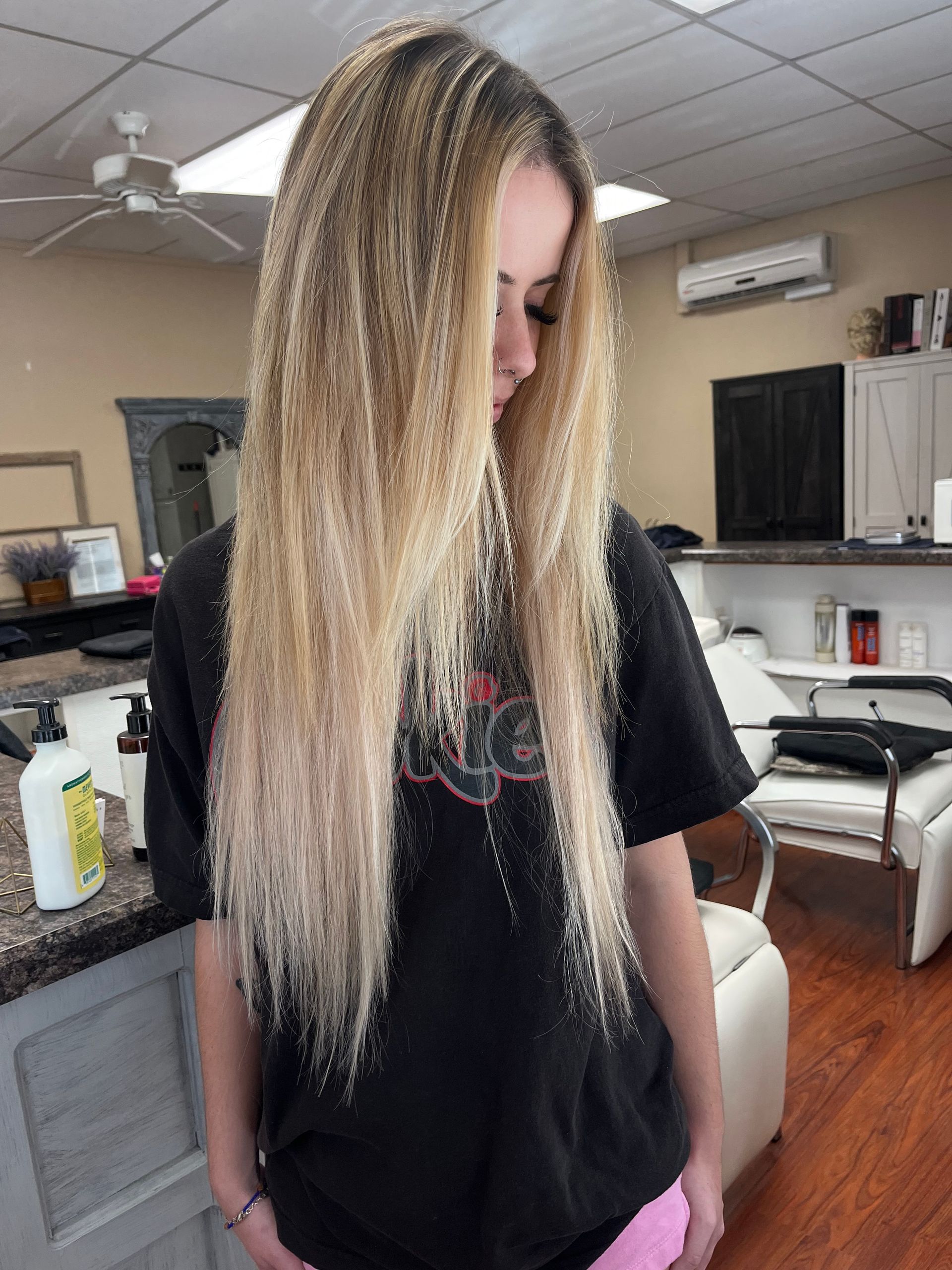A young woman with long blonde hair stands in a salon. She wears a black t-shirt, and the background includes salon equipment.