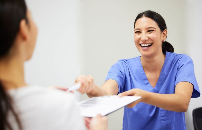 Smiling person in scrubs hands paperwork to another person in a light-filled office.