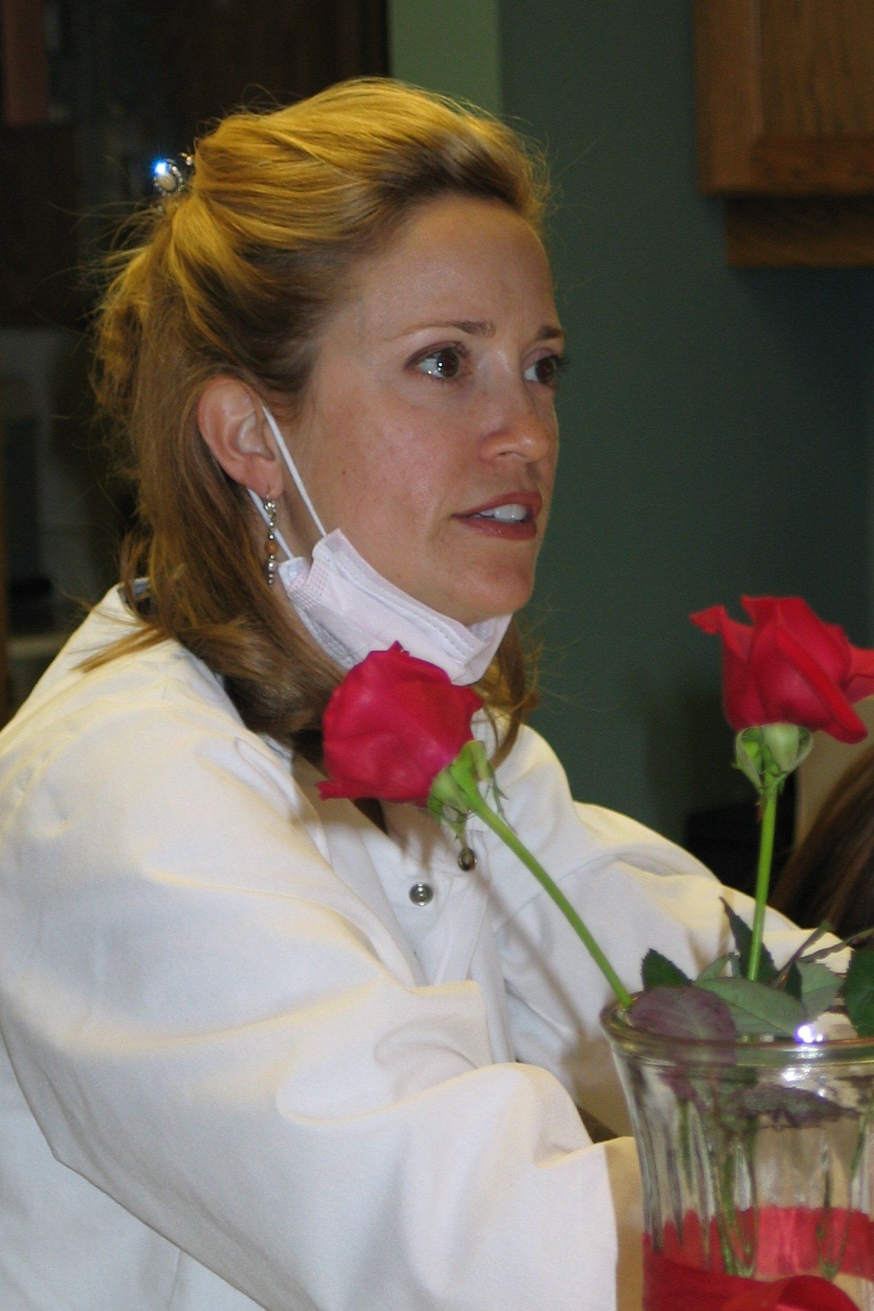 A woman wearing a mask and a white coat in front of a vase of red roses
