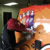 Person wrapping a colorful mural on an orange van side in a garage