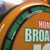 Close-up of an orange and black business sign with green text reading “Power 92.1 FM” and “Home Broad…”