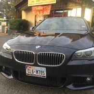 Black BMW sedan parked on a street at dusk, front view with headlights and storefront lights behind it