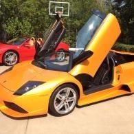 Orange sports car with scissor doors open, parked outdoors near a basketball hoop and a red car.