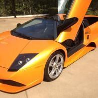 Orange sports car with gullwing door open, parked outdoors in sunlight