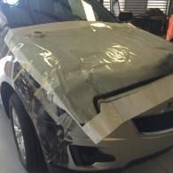 Damaged beige car with a crumpled hood in a garage, covered partly with clear plastic wrap