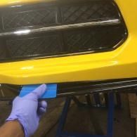 Gloved hand applying blue tape to a yellow car bumper near a black grille