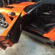 Orange sports car with driver’s door open in a garage, showing black interior and rear wheel.