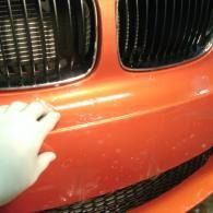Hand touching an orange car bumper below a black grille, showing a small dent or scratch