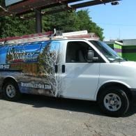 White utility van with a large wildlife-themed side graphic parked outdoors under a metal awning.