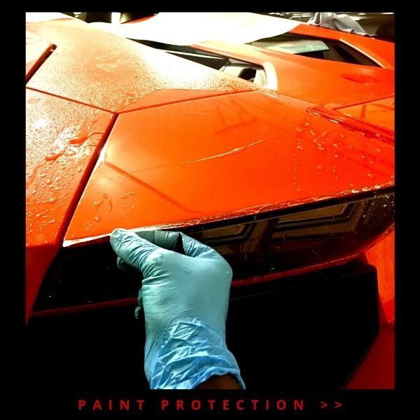 Blue-gloved hand applying paint protection film to an orange sports car hood