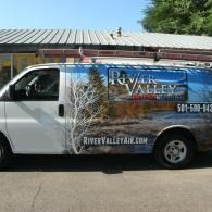 White van with River Valley landscaping wrap parked outdoors, featuring blue graphics and tree imagery.
