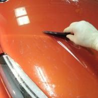 Gloved hand scraping debris off a glossy orange car hood with a tool