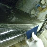A hand uses a blue applicator to smooth a black car panel in a garage.