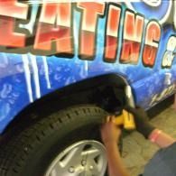 Worker using a power tool on a blue car with red lettering, near the front wheel
