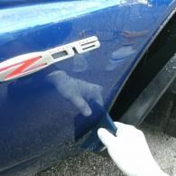 Gloved hand pointing at a dent on a blue car door near a Z06 badge