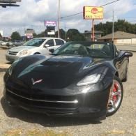 Black Chevrolet Corvette parked in a lot, with a white SUV beside it and roadside signs in the background
