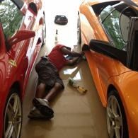 Person lying between parked cars, reaching toward an orange car in a narrow garage.