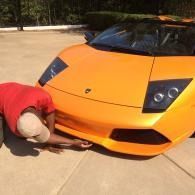 Person in a red shirt crouches under the front of an orange sports car on a driveway.
