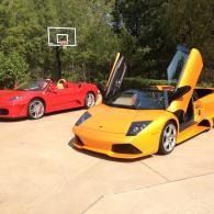 Two sports cars on a driveway, a yellow Lamborghini with scissor doors open and a red Ferrari beside it.