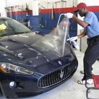 Man in a garage peeling protective film from the hood of a dark blue Maserati