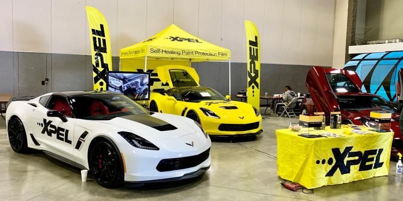 Three XPEL-branded sports cars on display at an auto show booth, including a white car, yellow car, and red car.