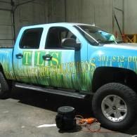 Blue and green lifted pickup truck with TLS branding parked in a garage, with tools on the floor.