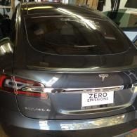Gray Tesla sedan in a garage, rear view with a “ZERO EMISSIONS” plate.