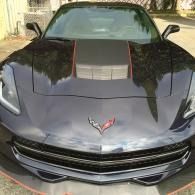 Front view of a black Corvette with a hood vent and red emblem, parked outdoors in daylight