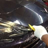 Hand wearing a white glove cleaning a black car hood with a yellow tool