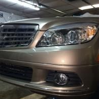 Front view of a beige sedan, showing the grille, headlight, and fog light in a garage.