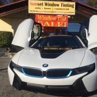 White BMW i8 with butterfly doors open in front of a Sunset Window Tinting sale sign