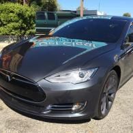 Dark gray Tesla sedan parked outdoors in sunlight, front view.