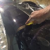 Hand scraping wet black car paint with a yellow-edged tool during detailing