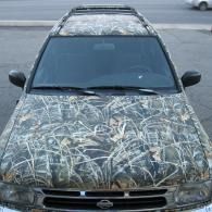 Car covered in camouflaged wrap, parked on pavement, front view.