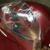 Close-up of a red car headlight covered in clear protective film, with a hand holding the edge