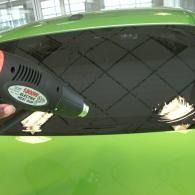 Heat gun heating the rear window of a green car, with a hand holding the tool.