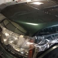Dark green car front with a damaged headlight wrapped in clear plastic.