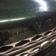 Close-up of a dark green car hood and grille with white paint splatters and water droplets