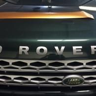 Close-up of a dark green Land Rover grille with silver ROVER letters and orange hood detail